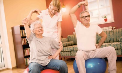 Senior caucasian patients sitting on an inflatable balloon while stretching their pronated arms and flexed wrist with the help of a young female physiotherapist